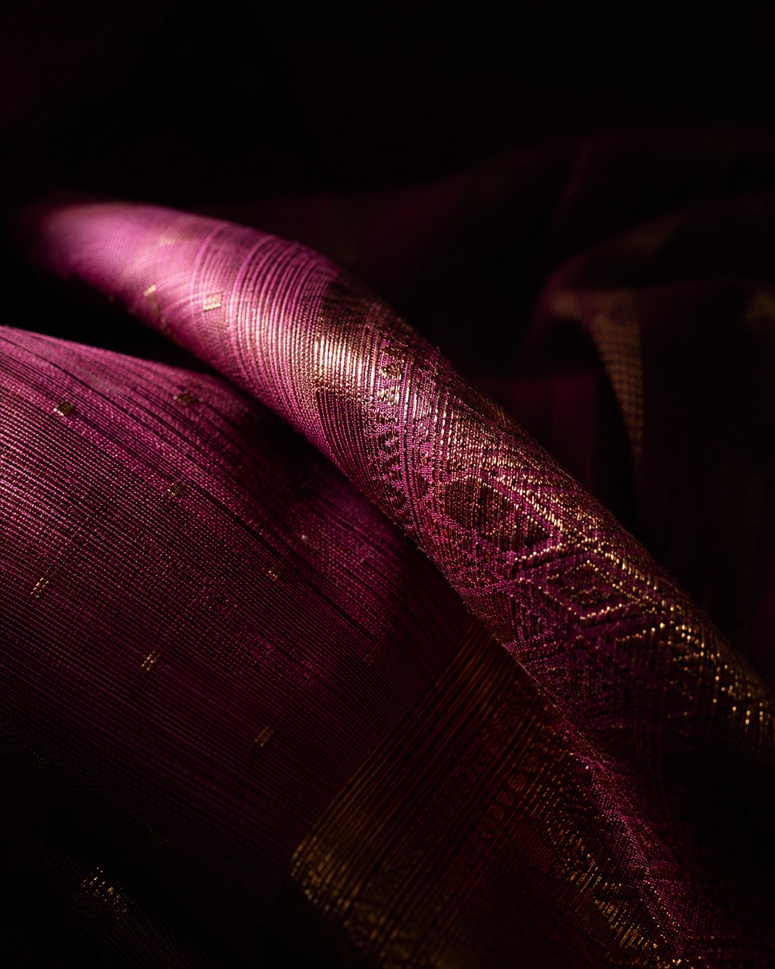 Close-up of deep magenta silk with gold thread woven through it, folds catching a single shaft of light.